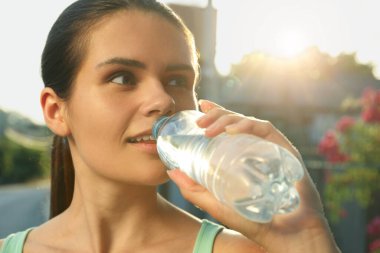 Happy young woman drinking water outdoors on hot summer day. Refreshing drink