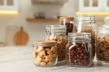 Glass containers with different breakfast cereals on white marble table in kitchen. Space for text