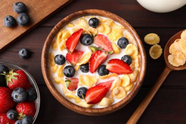 Bowl of tasty crispy corn flakes with milk and berries on wooden table, flat lay