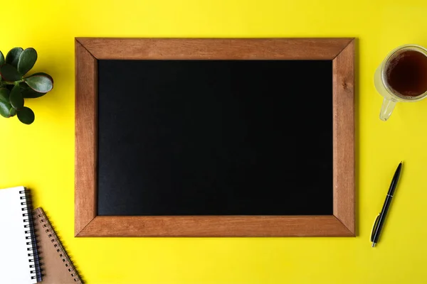 Clean small chalkboard, coffee, plant and stationery on yellow background, flat lay