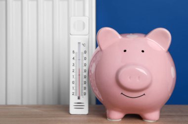 Piggy bank with thermometer on wooden table near heating radiator. Space for text