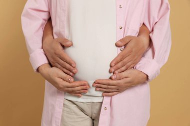 Man hugging his pregnant wife on light brown background, closeup