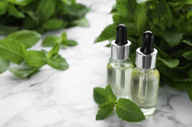 Bottles of mint essential oil and green leaves on white marble table, space for text