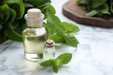 Bottle of mint essential oil and green leaves on white marble table, space for text