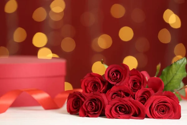 Beautiful red roses and gift box on white table against blurred lights, space for text. St. Valentine\'s day celebration