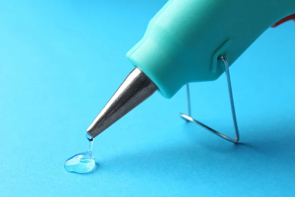 Melted glue dripping out of hot gun nozzle on light blue background, closeup