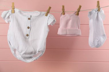Bebek kıyafetleri ve aksesuarlar pembe ahşap duvarın yanındaki çamaşır ipinde asılı.
