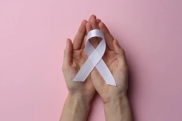 Woman holding white awareness ribbon on pink background, top view
