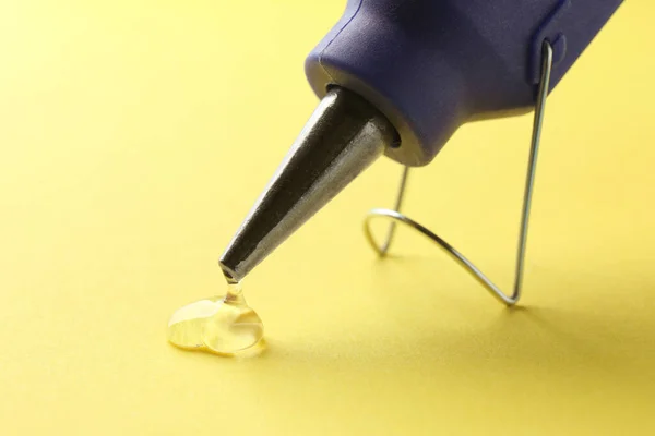 Melted glue dripping out of hot gun nozzle on yellow background, closeup