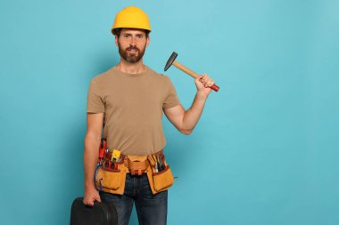 Professional builder in hard hat with hammer and tool belt on light blue background, space for text