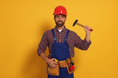 Professional builder in uniform with hammer and tool belt on yellow background
