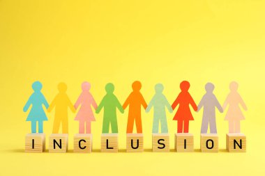 Paper human figures and wooden cubes with word Inclusion on yellow background