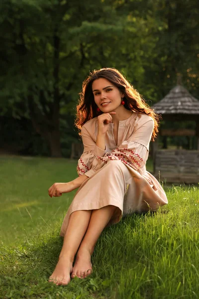 Beautiful woman in embroidered dress sitting on green grass in village. Ukrainian national clothes