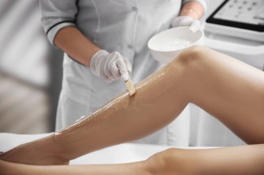 Professional cosmetologist applying gel on client's leg before laser epilation procedure in salon, closeup
