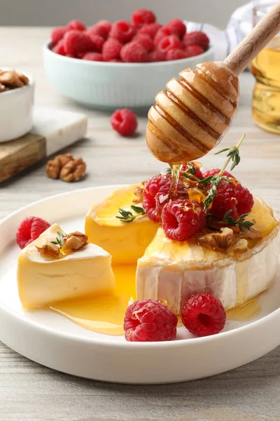 Pouring honey onto brie cheese served with raspberries and walnuts on white wooden table, closeup