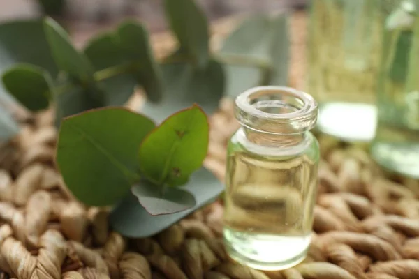 Bottle of eucalyptus essential oil and plant branches on wicker mat, closeup