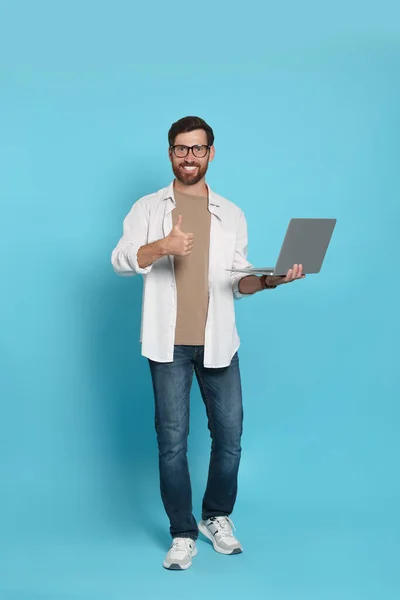 Handsome man with laptop showing thumb up gesture on light blue background