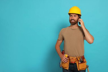 Professional builder in hard hat with tool belt talking on phone against light blue background, space for text