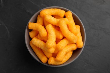 Many tasty cheesy corn puffs in bowl on black table, top view