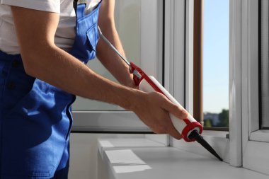 Worker sealing plastic window with caulk indoors, closeup. Installation process