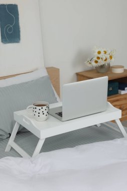 Laptoplu beyaz tepsi masası ve yatağın içinde bir bardak içki.