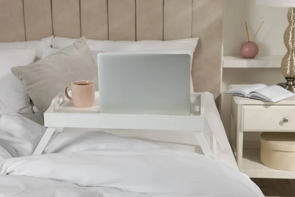 White tray table with laptop and cup of drink on bed indoors