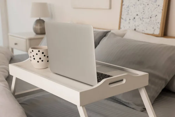 White tray table with laptop and cup of drink on bed indoors