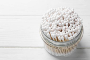Many cotton buds in glass jar on white wooden table, space for text