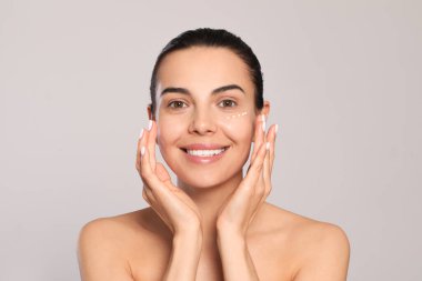 Beautiful young woman with cream on skin under eye against light grey background