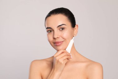 Beautiful young woman with cream on skin under eye against light grey background