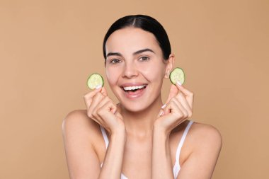 Beautiful young woman with slices of cucumber on beige background