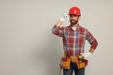 Professional builder in hard hat with tool belt on light background, space for text