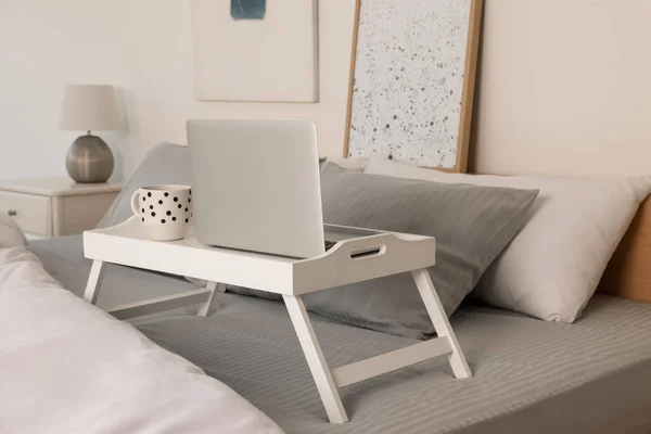 White tray table with laptop and cup of drink on bed indoors