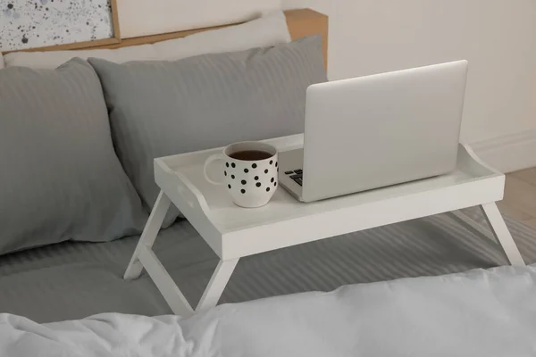 White tray table with laptop and cup of drink on bed indoors