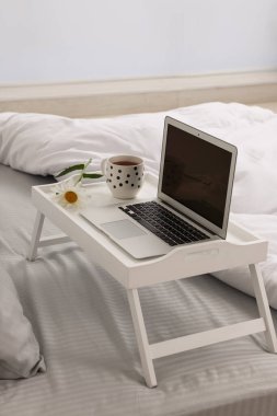 White tray table with laptop, cup of drink and daisy on bed indoors
