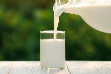 Pouring tasty fresh milk from jug into glass on white wooden table, closeup
