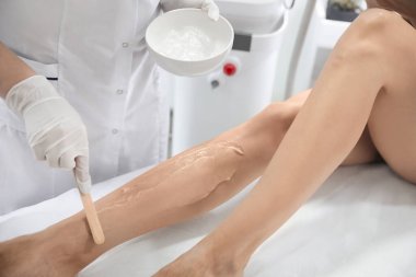 Professional cosmetologist applying gel on client's leg before laser epilation procedure in salon, closeup