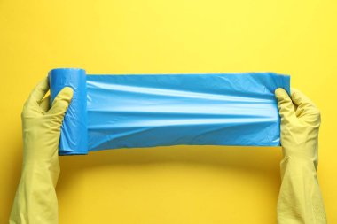 Janitor in rubber gloves holding roll of light blue garbage bags over yellow background, top view