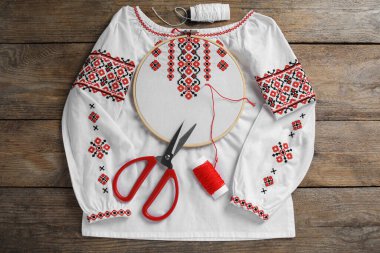 Shirt with red embroidery design in hoop, needle, scissors and threads on wooden table, flat lay. National Ukrainian clothes