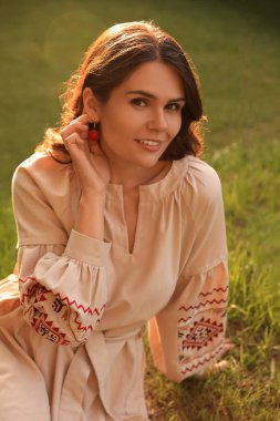 Beautiful woman in embroidered dress sitting on green grass outdoors. Ukrainian national clothes