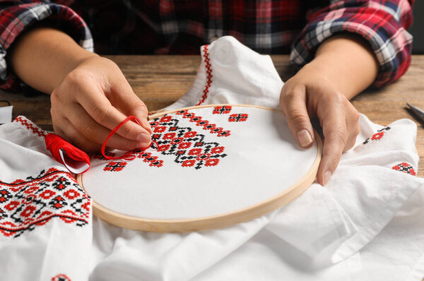 Woman embroidering white shirt with colorful threads at wooden table, closeup. Ukrainian national clothes