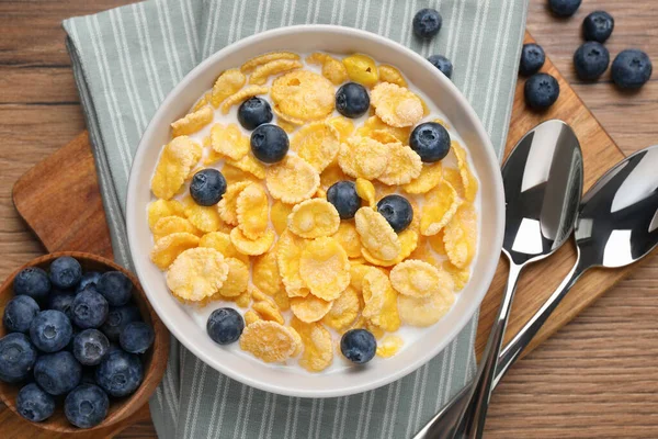 Bowl of tasty crispy corn flakes with milk and blueberries on wooden table, flat lay