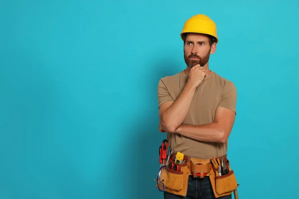 Professional builder in hard hat with tool belt on light blue background, space for text