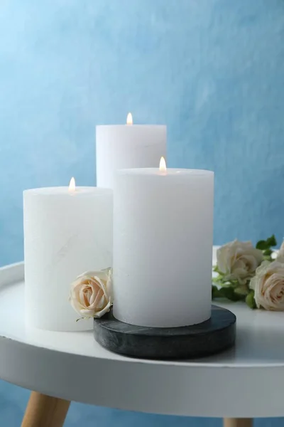 Burning candles and beautiful roses on white table near light blue wall