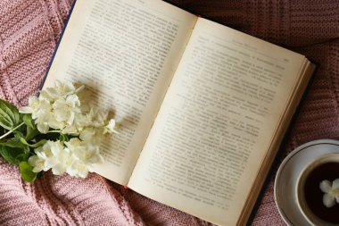 Beautiful jasmine flowers, open book and cup aromatic tea on pink fabric, flat lay