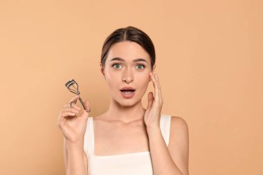 Emotional woman with eyelash curler on beige background