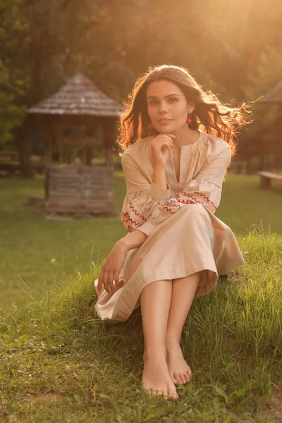 Beautiful woman in embroidered dress sitting on green grass in village. Ukrainian national clothes
