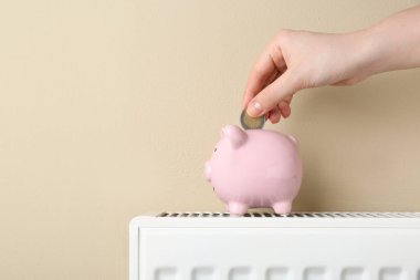 Woman putting coin into piggy bank on heating radiator against beige background, closeup. Space for text