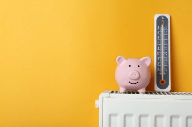Piggy bank and thermometer on heating radiator against orange background, space for text