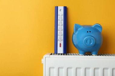 Piggy bank and thermometer on heating radiator against orange background, space for text